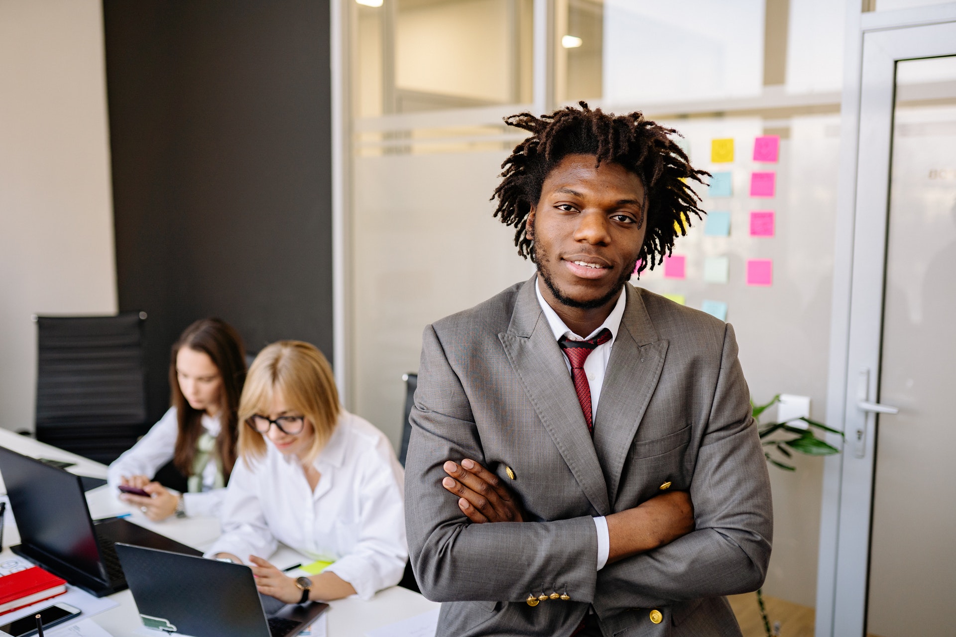 Black Hr professional man with arms crossed and female colleagues discussing HR functions behind him